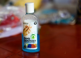 A bottle of hand sanitizer is placed on a reddish-brown surface. The label on the bottle includes an image of hands and text indicating it is 100ml and provides protection from germs. The background is blurred, with indistinct objects and a mix of colors.