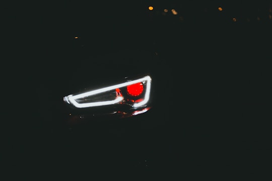 A close-up view of a car headlight in low light conditions with the red and white lights prominently glowing against a dark background.