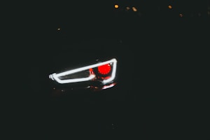 A close-up view of a car headlight in low light conditions with the red and white lights prominently glowing against a dark background.