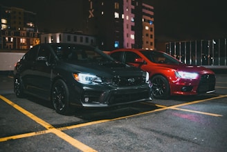 Two cars are parked side by side in an outdoor parking lot during nighttime. The car on the left is black, with illuminated headlights creating a contrast against the dark environment. The car on the right is red, also showcasing bright headlights. Surrounding buildings and glowing windows provide an urban backdrop.