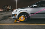 A silver car is parked in a lot with a wheel lock, also known as a boot, attached to its front tire. The lock is yellow with a red warning sign and is secured with a pink strap. The car is slightly rusted near the wheel well. In the background, a multi-story building is visible with lights illuminating through its windows. The parking area is marked with a yellow line and the number '52' is painted on the ground.