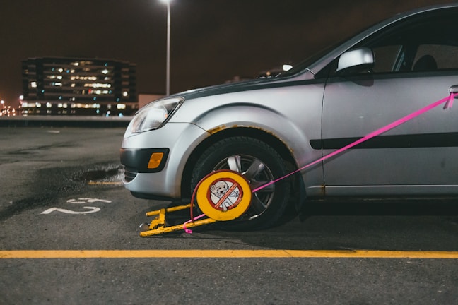 A silver car is parked in a lot with a wheel lock, also known as a boot, attached to its front tire. The lock is yellow with a red warning sign and is secured with a pink strap. The car is slightly rusted near the wheel well. In the background, a multi-story building is visible with lights illuminating through its windows. The parking area is marked with a yellow line and the number '52' is painted on the ground.