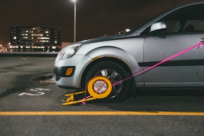 A silver car is parked in a lot with a wheel lock, also known as a boot, attached to its front tire. The lock is yellow with a red warning sign and is secured with a pink strap. The car is slightly rusted near the wheel well. In the background, a multi-story building is visible with lights illuminating through its windows. The parking area is marked with a yellow line and the number '52' is painted on the ground.