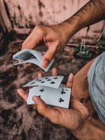 A person skillfully shuffles a deck of playing cards with both hands. The cards are captured in mid-motion, showing a variety of suits and numbers. The background appears to be an outdoor setting with a textured surface.