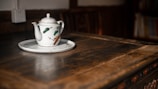 A white ceramic teapot with floral design sits on a matching saucer, placed on a rustic wooden table. The warm light creates a cozy atmosphere, gently highlighting the details of the teapot and table.