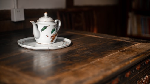 A white ceramic teapot with floral design sits on a matching saucer, placed on a rustic wooden table. The warm light creates a cozy atmosphere, gently highlighting the details of the teapot and table.