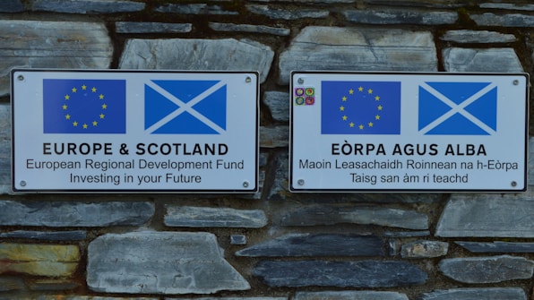 Two signs mounted on a stone wall display the European Union and Scottish flags. The left sign is in English stating 'EUROPE & SCOTLAND European Regional Development Fund Investing in your Future.' The right sign is in Scottish Gaelic, conveying the same message.