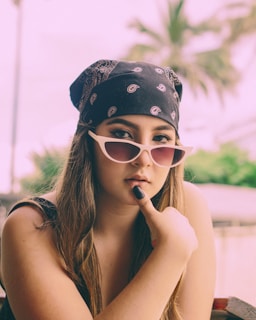 A young woman wearing a black bandana on her head and stylish white-framed sunglasses. She has long brown hair and is holding her index finger to her chin, possibly in a thoughtful pose. In the background, there are palm trees and a blurred outdoor setting with green foliage.