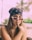 A young woman wearing a black bandana on her head and stylish white-framed sunglasses. She has long brown hair and is holding her index finger to her chin, possibly in a thoughtful pose. In the background, there are palm trees and a blurred outdoor setting with green foliage.