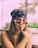 A young woman wearing a black bandana on her head and stylish white-framed sunglasses. She has long brown hair and is holding her index finger to her chin, possibly in a thoughtful pose. In the background, there are palm trees and a blurred outdoor setting with green foliage.