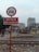 A railway crossing area with a sign that has a no-car symbol and Vietnamese text. There are railway tracks in the foreground, a construction vehicle, and a few buildings in the background. People on motorbikes are visible, and the overall setting appears to be urban with overcast weather.