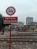 A railway crossing area with a sign that has a no-car symbol and Vietnamese text. There are railway tracks in the foreground, a construction vehicle, and a few buildings in the background. People on motorbikes are visible, and the overall setting appears to be urban with overcast weather.