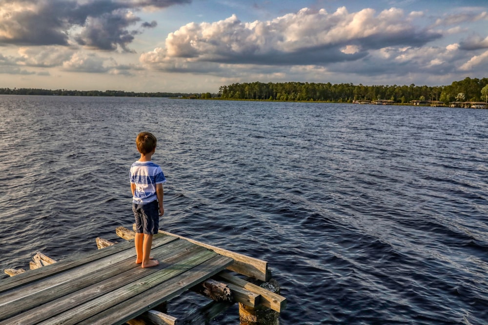boy in white t-shirt standing on wooden dock during daytime