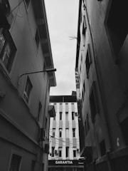 A black and white photograph of a narrow alleyway between two tall buildings. The buildings show visible signs of wear, with windows and some air conditioning units. At the end of the alley, another structure with vertical lines is visible, with the word 'GARANTIA' on a sign.