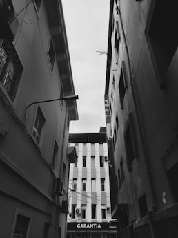 A black and white photograph of a narrow alleyway between two tall buildings. The buildings show visible signs of wear, with windows and some air conditioning units. At the end of the alley, another structure with vertical lines is visible, with the word 'GARANTIA' on a sign.
