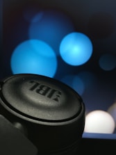 A close-up view of a black JBL headphone with a blurred background featuring large blue bokeh circles.