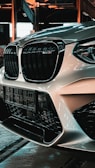 Close-up view of a car's front end, featuring a prominent black grille and headlight design. The car is parked in an industrial setting with exposed beams and large windows. The light reflects off the car's glossy surface, creating a sleek and polished appearance.
