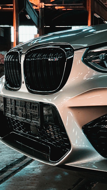 Close-up view of a car's front end, featuring a prominent black grille and headlight design. The car is parked in an industrial setting with exposed beams and large windows. The light reflects off the car's glossy surface, creating a sleek and polished appearance.