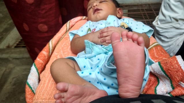 A baby is peacefully sleeping while lying on a striped orange towel, wearing a light blue outfit with small blue polka dots. The baby's hands are folded on their chest, and a black bracelet is visible on the wrist. The focus is on the baby's feet, with one foot prominently in the foreground.
