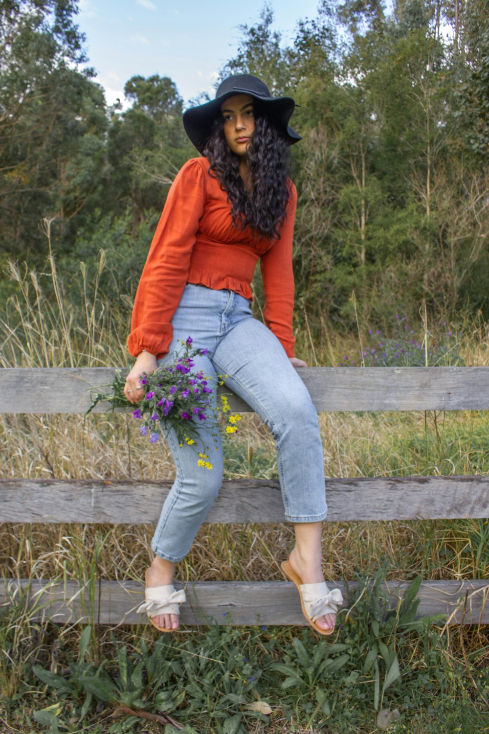 woman in red long sleeve shirt and blue denim jeans sitting on brown wooden fence during