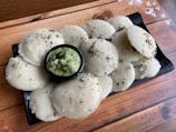 A plate filled with multiple white, round steamed rice cakes garnished with mustard seeds, accompanied by a small bowl of green chutney topped with curry leaves. The background features a wooden surface with a subtle pattern of white flowers.