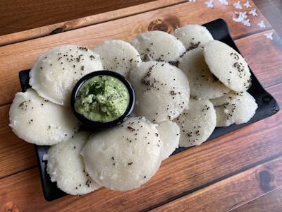 A plate filled with multiple white, round steamed rice cakes garnished with mustard seeds, accompanied by a small bowl of green chutney topped with curry leaves. The background features a wooden surface with a subtle pattern of white flowers.