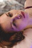 A person is lying down with a serene expression, illuminated by a soft, colorful light that casts purple and pink hues on their face. The background is blurred, creating a gentle atmosphere.