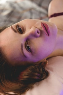 A person is lying down with a serene expression, illuminated by a soft, colorful light that casts purple and pink hues on their face. The background is blurred, creating a gentle atmosphere.