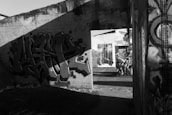 A black and white photograph of an urban setting with graffiti art on concrete walls. The scene includes several pieces of street art, featuring abstract designs and figures. Shadows cast across the walls, adding dramatic contrast to the artwork. The environment appears to be an abandoned or neglected area, suggesting a sense of decay.