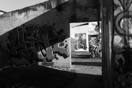 A black and white photograph of an urban setting with graffiti art on concrete walls. The scene includes several pieces of street art, featuring abstract designs and figures. Shadows cast across the walls, adding dramatic contrast to the artwork. The environment appears to be an abandoned or neglected area, suggesting a sense of decay.
