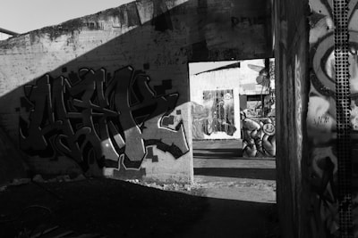A black and white photograph of an urban setting with graffiti art on concrete walls. The scene includes several pieces of street art, featuring abstract designs and figures. Shadows cast across the walls, adding dramatic contrast to the artwork. The environment appears to be an abandoned or neglected area, suggesting a sense of decay.