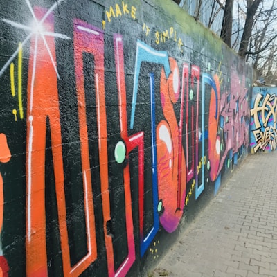 A vibrant graffiti mural on a concrete wall, featuring abstract and geometric shapes in bold colors like orange, pink, and blue. The words 'MAKE IT SIMPLE' are prominently displayed at the top. The artwork extends along the wall, accompanied by dynamic patterns and designs.