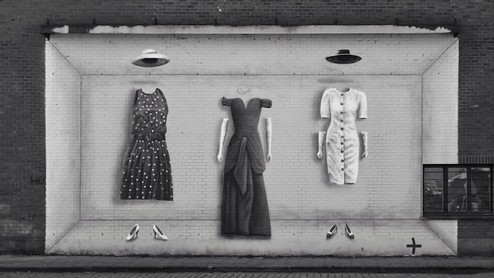 A black and white mural on a brick wall depicts three dresses with matching hats and shoes arranged in a row. The dresses appear to be three-dimensional and are painted to mimic clothing hanging as if on display. The left dress has polka dots, the middle dress is long with structured sleeves, and the right dress features buttons down the front. Below each dress is a pair of shoes, and above are hats suspended in the air.