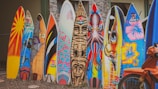 A row of colorful surfboards stands upright on a gravel surface. Each surfboard is uniquely painted with various vibrant designs and illustrations, including patterns, abstract art, and depictions of animals. In the foreground, part of an orange bicycle is visible.