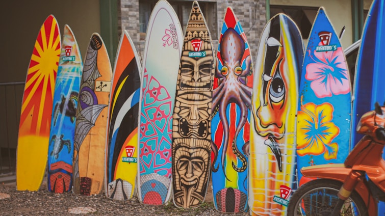A row of colorful surfboards stands upright on a gravel surface. Each surfboard is uniquely painted with various vibrant designs and illustrations, including patterns, abstract art, and depictions of animals. In the foreground, part of an orange bicycle is visible.