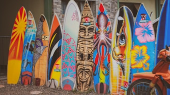 A row of colorful surfboards stands upright on a gravel surface. Each surfboard is uniquely painted with various vibrant designs and illustrations, including patterns, abstract art, and depictions of animals. In the foreground, part of an orange bicycle is visible.