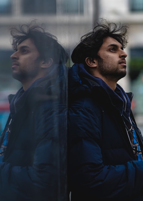 A person is seen in profile, wearing a dark jacket and leaning against a reflective glass surface, creating a mirror image effect. The background appears to be urban, slightly blurred, giving a sense of depth. The lighting is subdued, emphasizing the introspective mood.
