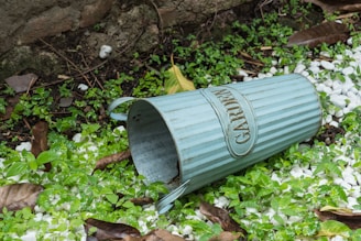 A metal container labeled 'GARDEN' lies on its side on the ground surrounded by green plants, white pebbles, and dried leaves. The background features some ferns and a stone wall.