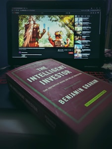 A laptop displaying a YouTube video featuring two animated characters holding popsicles is placed in the background. In the foreground, the book 'The Intelligent Investor' by Benjamin Graham is prominently displayed on a surface.