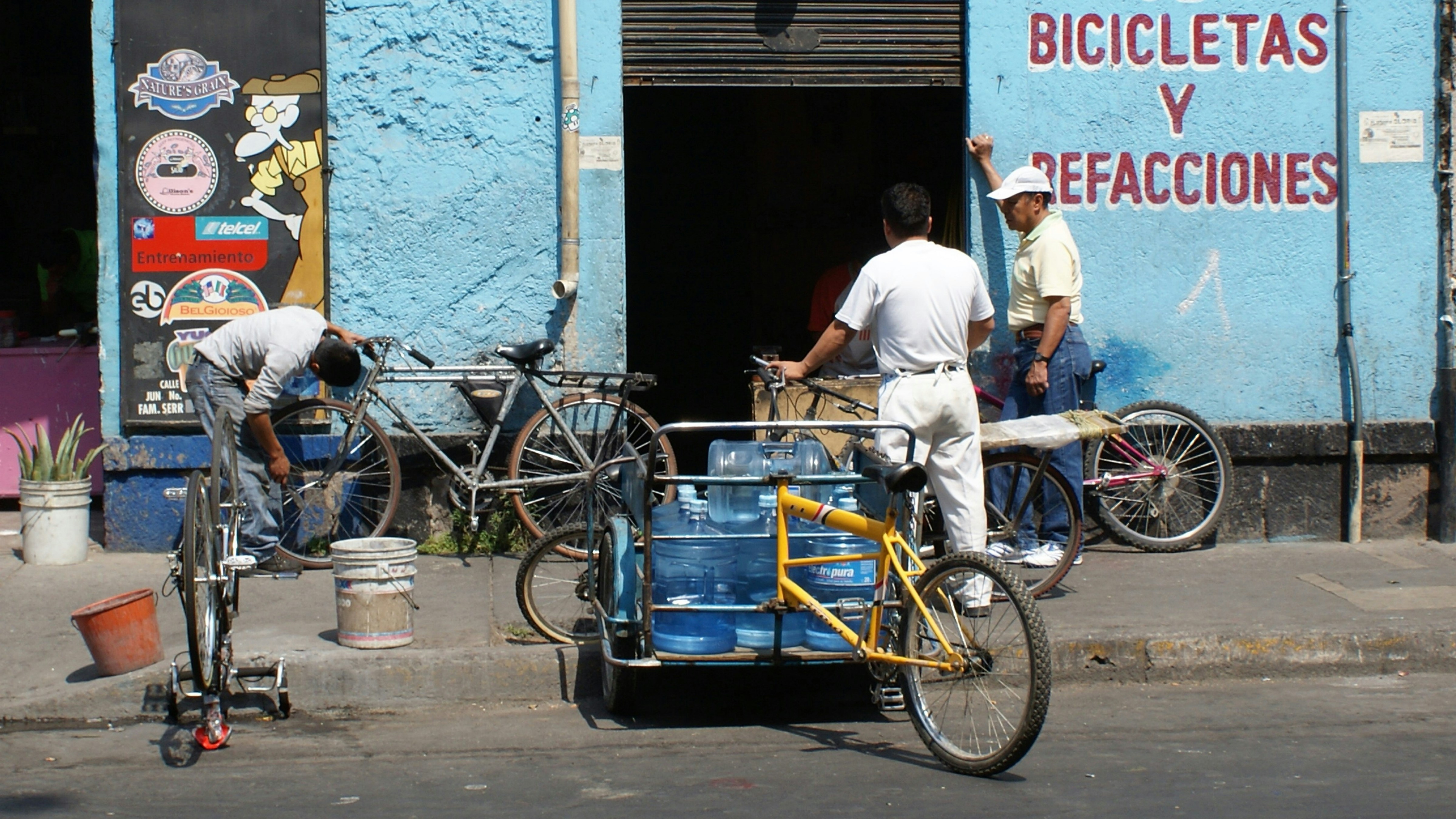 A scene in front of a bicycle repair shop with three men and several bicycles. One man is working on a bike while another leans against the wall, engaging in conversation. A tricycle with blue water containers also occupies the foreground. The building is painted light blue with signage in red lettering.