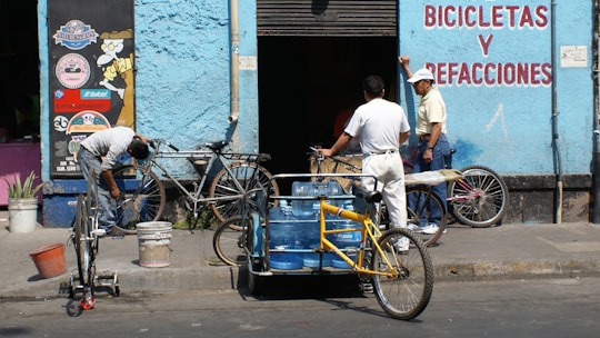 A scene in front of a bicycle repair shop with three men and several bicycles. One man is working on a bike while another leans against the wall, engaging in conversation. A tricycle with blue water containers also occupies the foreground. The building is painted light blue with signage in red lettering.