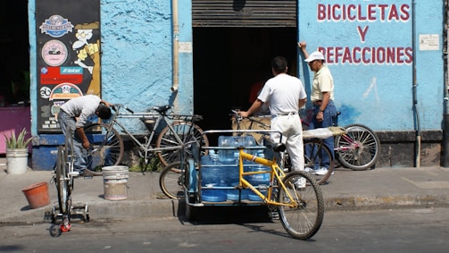 A scene in front of a bicycle repair shop with three men and several bicycles. One man is working on a bike while another leans against the wall, engaging in conversation. A tricycle with blue water containers also occupies the foreground. The building is painted light blue with signage in red lettering.