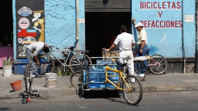 A scene in front of a bicycle repair shop with three men and several bicycles. One man is working on a bike while another leans against the wall, engaging in conversation. A tricycle with blue water containers also occupies the foreground. The building is painted light blue with signage in red lettering.