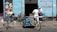 A scene in front of a bicycle repair shop with three men and several bicycles. One man is working on a bike while another leans against the wall, engaging in conversation. A tricycle with blue water containers also occupies the foreground. The building is painted light blue with signage in red lettering.