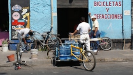 A scene in front of a bicycle repair shop with three men and several bicycles. One man is working on a bike while another leans against the wall, engaging in conversation. A tricycle with blue water containers also occupies the foreground. The building is painted light blue with signage in red lettering.