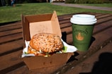 A sesame seed burger in a cardboard takeout box sits on a wooden table outdoors, with a green paper cup labeled Campos beside it, against a backdrop of green grass.