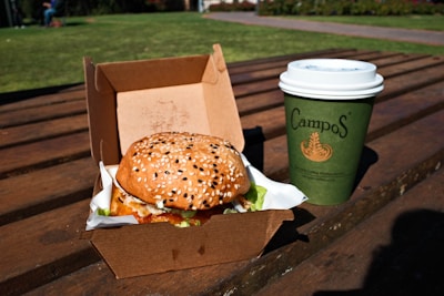 A sesame seed burger in a cardboard takeout box sits on a wooden table outdoors, with a green paper cup labeled Campos beside it, against a backdrop of green grass.