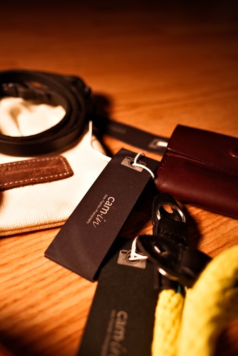 Various leather and fabric accessories are arranged on a wooden surface under warm lighting. Tags with branding are attached to some items, featuring text like 'cam'in'. A coiled belt, a leather pouch, and a braided yellow cord or strap are visible.