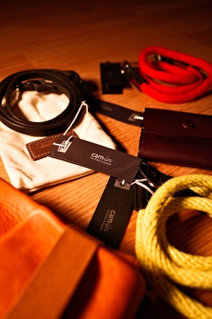 A collection of fashion accessories, including belts and bracelets, is artistically arranged on a wooden surface. The items feature various materials and colors, such as leather and fabric, with visible brand tags attached. Rich lighting adds warmth to the scene, highlighting the textures of each piece.