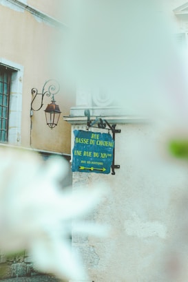 A metal street sign in French is affixed to the side of a light-colored building. The sign reads 'Rue Basse Du Chateau' and directs to 'Une Rue Du XIVème' with two arrows pointing in opposite directions for shops. It has a blue background with yellow text. Next to the sign, there is an ornate metal bracket holding an old-fashioned lantern. The background includes window frames and other architectural features of a classical building. In the foreground, there are blurred white flowers, likely indicating a shallow depth of field in the photograph.